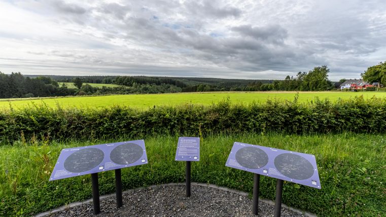 Eine Aussichtsplattform mit Informationstafeln steht vor einer grünen Landschaft. Der Himmel ist bewölkt und es gibt eine Weitsicht auf die Felder.