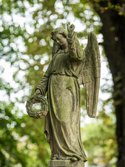 Eine Statue eines Engels mit Flügeln und einer Kranz in der Hand. Die Statue steht umgeben von grünen Bäumen.