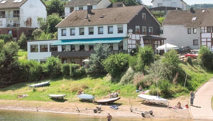 Ein malerisches Haus am Ufer mit Booten und einer schönen Wiese. Menschen entspannen sich am Strand und genießen die Sonne.
