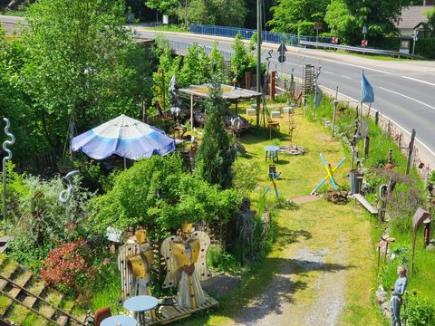 Ein origineller Garten mit verschiedenen Skulpturen und einem großen Sonnenschirm. Die grüne Umgebung und der Weg führen zu einer Straße.
