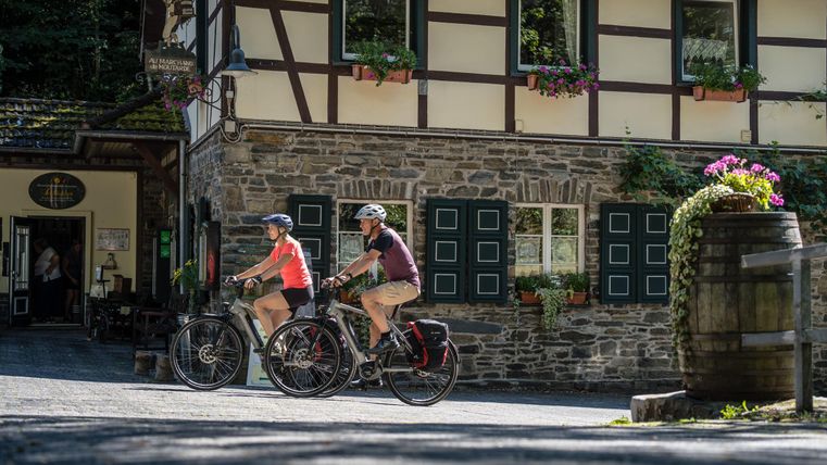Zwei Radfahrer fahren an einem rustikalen Gebäude mit Blumenfenstern vorbei. Die Sonne scheint und schafft eine einladende Atmosphäre im Freien.