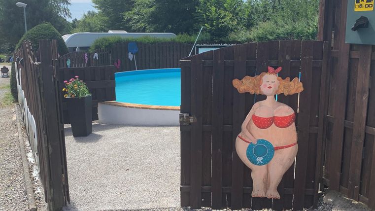 Ein Eingang zu einem Garten mit einem blauen Pool und einer bunten Wandmalerei einer fröhlichen Frau in einem roten Badeanzug. Der Bereich ist gut gepflegt und hat Blumen in einem Topf.