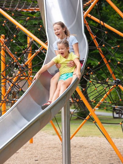 Eine Mutter und ihr Kind rutschen fröhlich von einer Rutsche. Im Hintergrund sind bunte Spielgeräte und ein grüner Park zu sehen.