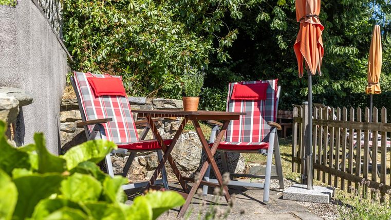 Ein einladender Gartenbereich mit zwei gestreiften Stühlen und einem kleinen Tisch. Im Hintergrund stehen Sonnenschirme und eine Holzlattenzaun umrahmt den Raum.
