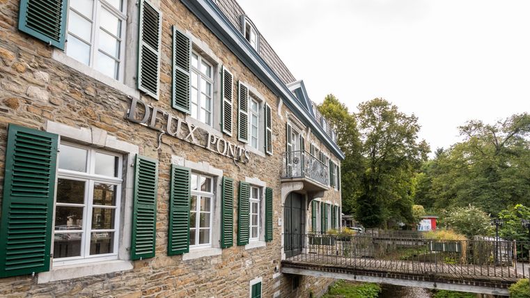 Ein historisches Gebäude mit grünen Fensterläden und einem Fluss im Vordergrund. Die Umgebung ist von Bäumen und Natur geprägt.