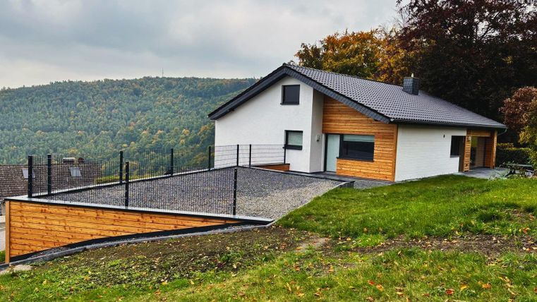 Ein modernes Haus mit einer Kombination aus Holz- und Putzfassade. Es liegt in einer grünen Umgebung mit sanften Hügeln im Hintergrund.