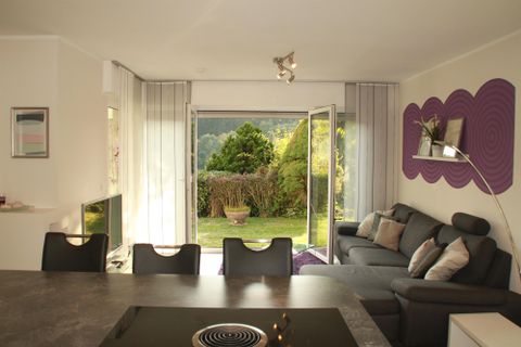 Gemütliches Wohnzimmer mit einer grauen Couch und einem Blick auf den Garten durch große Fenster. Der Raum hat eine moderne, helle Atmosphäre.