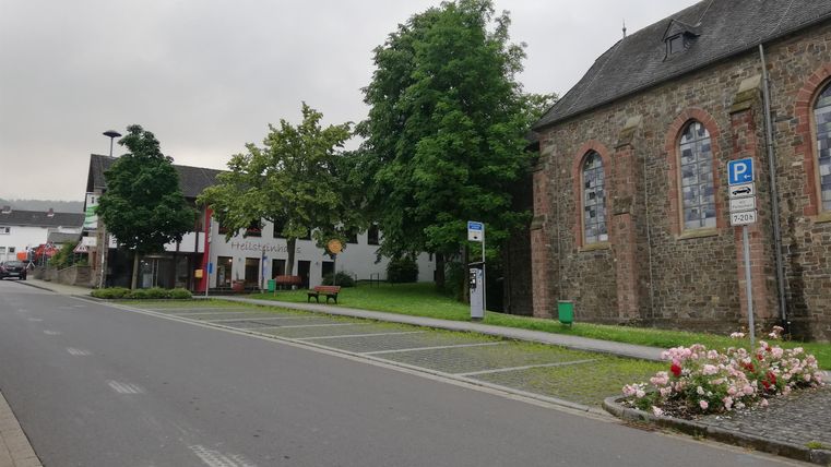 Eine ruhige Straße mit grünen Bäumen und einer alten Kirche. Links sind einige Gebäude und Blumenbeete zu sehen.