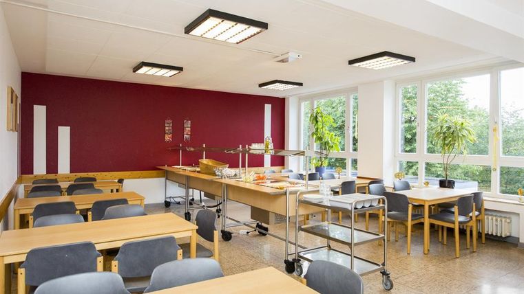 Ein helle Essensraum mit großen Fenstern und einem modernen Ambiente. Es gibt mehrere Tische mit Stühlen und eine Servierstation für Speisen.