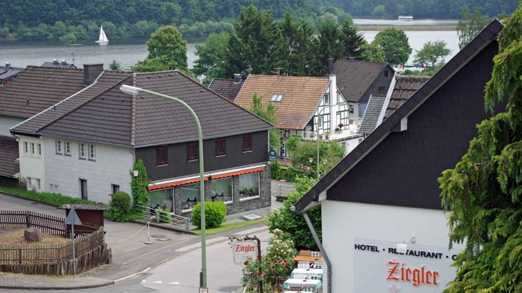 Eine malerische Aussicht auf ein kleines Dorf am Fluss. Im Vordergrund sind mehrere Häuser und ein Restaurant zu sehen, umgeben von grüner Natur.