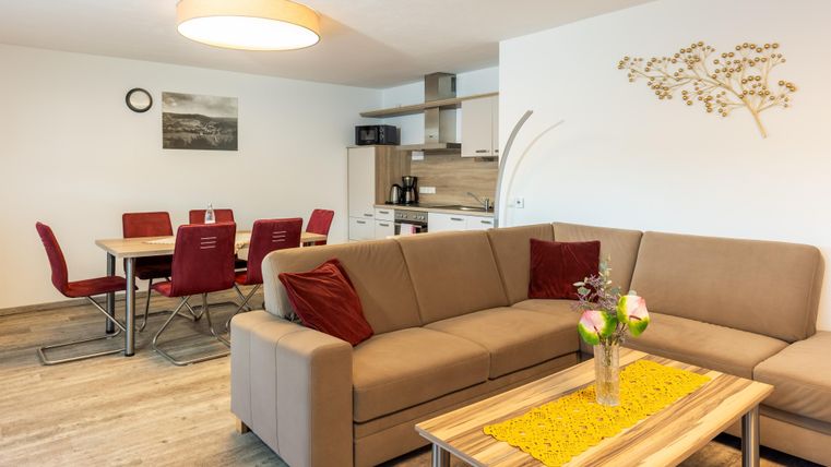 Ein modernes Wohnzimmer mit einem beige Sofa und roten Kissen. Im Hintergrund befindet sich eine offene Küche mit einem Essbereich.