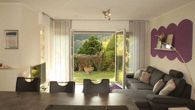 Gemütliches Wohnzimmer mit einer grauen Couch und einem Blick auf den Garten durch große Fenster. Der Raum hat eine moderne, helle Atmosphäre.