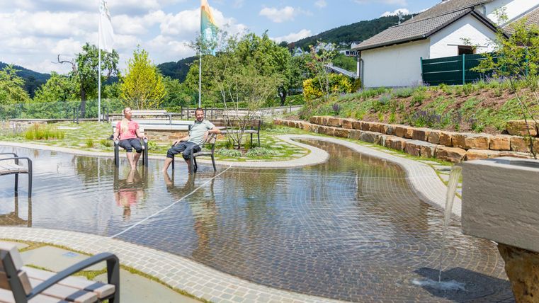 Ein ruhiger Garten mit einem Wasserbecken und zwei Personen, die entspannen. Umgeben von grünen Pflanzen und einem klaren Himmel.