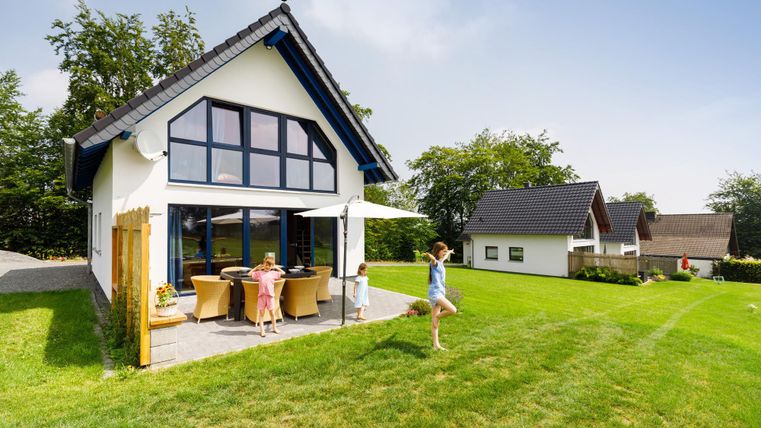 Ein modernes Haus mit großen Fenstern steht in einem grünen Garten. Kinder spielen auf dem Rasen und genießen die Sonne.