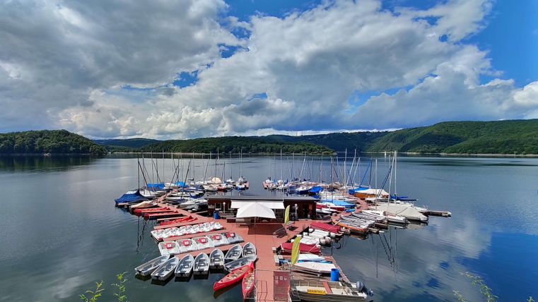 Ein Hafen mit mehreren Booten und Yachten liegt ruhig am Wasser. Im Hintergrund sind grüne Hügel und ein blauer Himmel mit Wolken zu sehen.