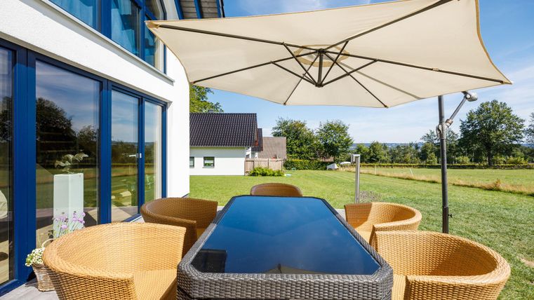 Ein schöner Esstisch im Freien mit Korbsesseln und einem Sonnenschirm. Die Umgebung ist grün und einladend.