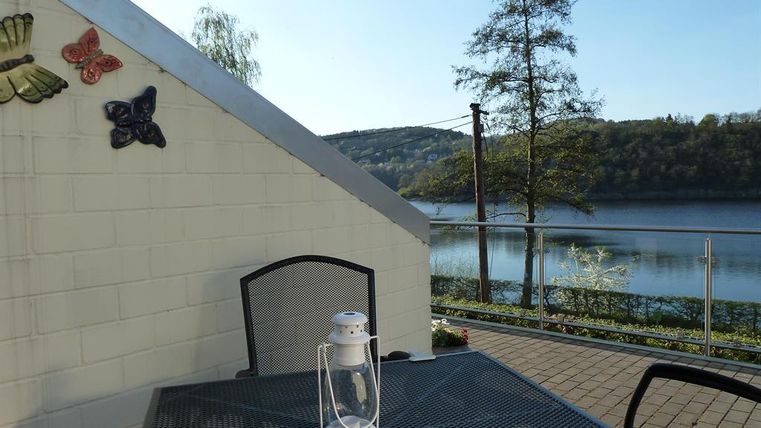 Ein gemütlicher Außenbereich mit einem Tisch und einer Laterne. Im Hintergrund ist ein ruhiger Fluss und grüne Bäume zu sehen.