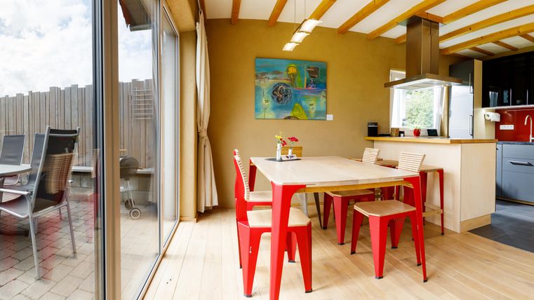 Eine helle Küche mit einem Esstisch und roten Stühlen. Durch große Fenster blickt man auf einen schönen Garten.