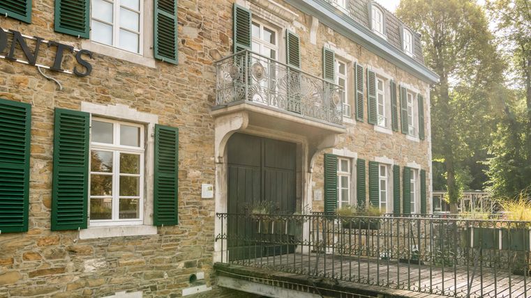 Ein historisches Gebäude aus Stein mit grünen Fensterläden und einem Balkon. Eine kleine Holzbrücke führt zum Eingang.