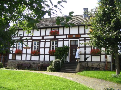 Ein traditionelles Fachwerkhaus mit blühenden Kästen an den Fenstern. Der Garten ist grün und einladend, und eine Person steht vor der Haustür.