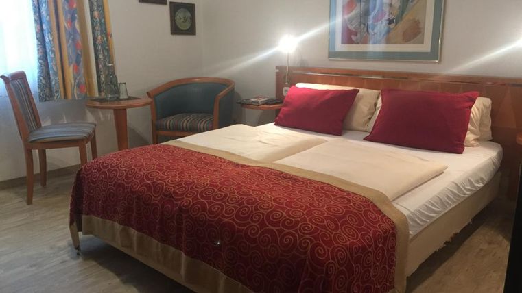 Ein gemütliches Schlafzimmer mit einem großen Bett und roten Kissen. Daneben stehen ein Tisch und ein Stuhl, sowie dekorative Bilder an der Wand.