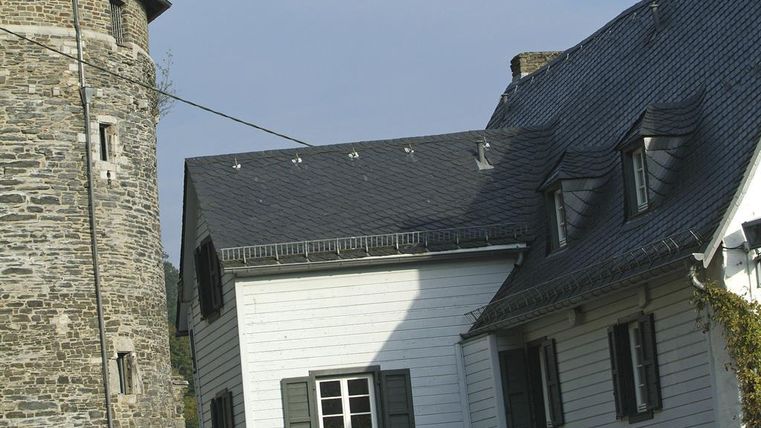 Ein historisches Gebäude mit einem steinernen Turm und einem weißen Haus. Das Dach des Hauses hat schöne Details und die Fenster sind mit grünen Fensterläden ausgestattet.