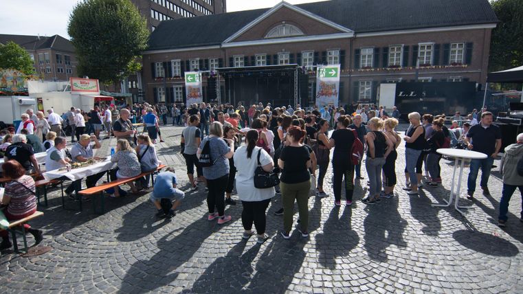 Eine große Menschenmenge versammelt sich auf einem Platz vor einem Gebäude. Im Hintergrund stehen Bühnen und Tische, während die Sonne scheint.