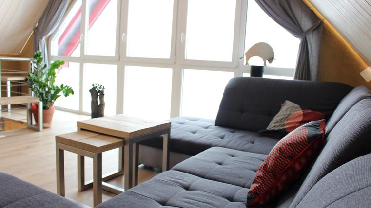 Ein gemütliches Wohnzimmer mit einem grauen Sofa und zwei kleinen Tischen. Durch große Fenster fällt viel Licht herein und sorgt für eine einladende Atmosphäre.