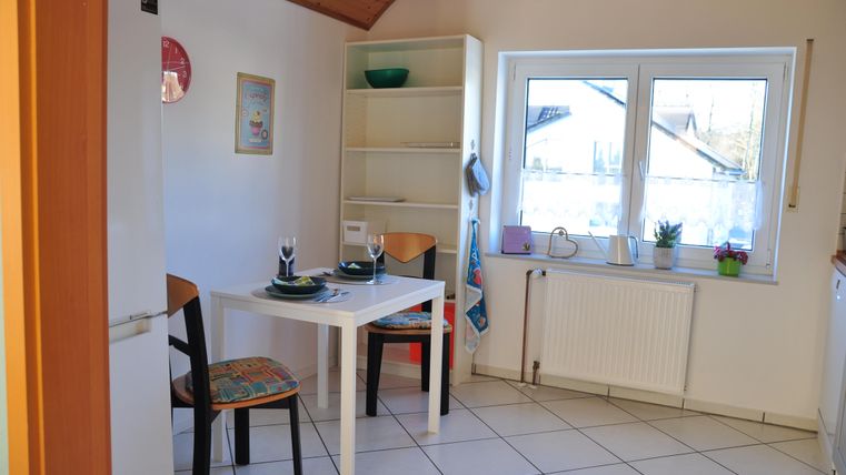 Eine helle Küche mit einem kleinen Tisch und zwei Stühlen. Im Hintergrund steht ein Regal und ein Fenster mit Blick nach draußen.