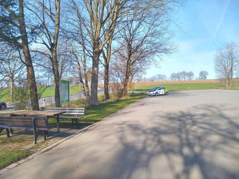 Ein ruhiger Parkplatz umgeben von Bäumen und Wiesen. Die Sonne scheint und der Himmel ist klar.