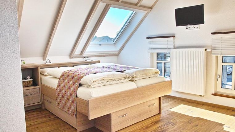 Ein gemütliches Schlafzimmer mit einem Doppelbett und Holzfußboden. Das Zimmer hat schräg verlaufende Wände, ein Fenster mit Ausblick und einen Fernseher an der Wand.