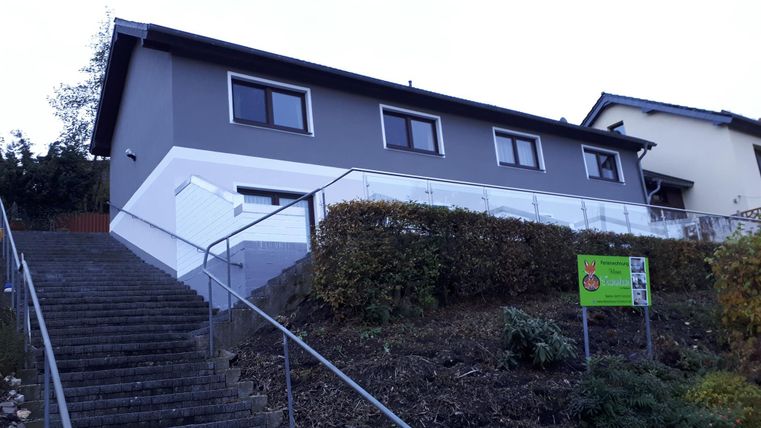 Ein modernes Gebäude mit grauer Fassade und großen Fenstern, umgeben von einem angelegten Bereich. Die Treppe führt zum Haupteingang des Hauses.