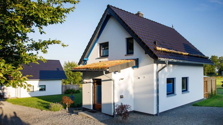 Ein modernes Einfamilienhaus mit einem Satteldach und blauem Fensterrahmen. Der Garten ist gepflegt, und die Umgebung ist grün und einladend.
