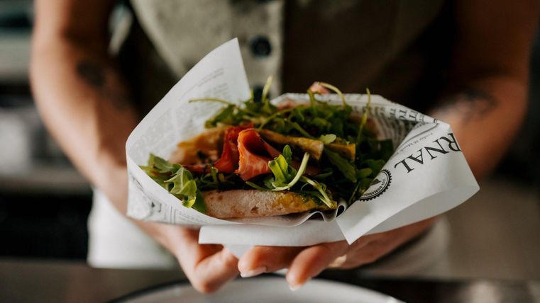 Ein frisch zubereitetes Sandwich mit Schinken und Salat, eingewickelt in Papier. Es wird von einer Person gehalten, die an einem Tisch steht.