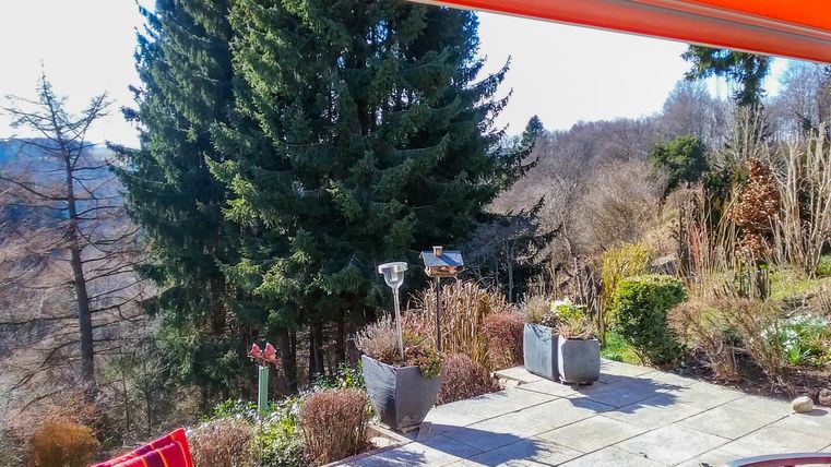 Eine Terrasse mit roten Stühlen und Blick auf eine bewaldete Landschaft. Die Sonne scheint und der Himmel ist blau.