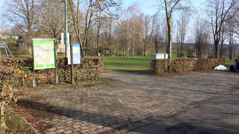 Ein Parkplatz, der an einen Park grenzt, mit Bänken und Schildern. Im Hintergrund sind Bäume und ein klarer Himmel zu sehen.