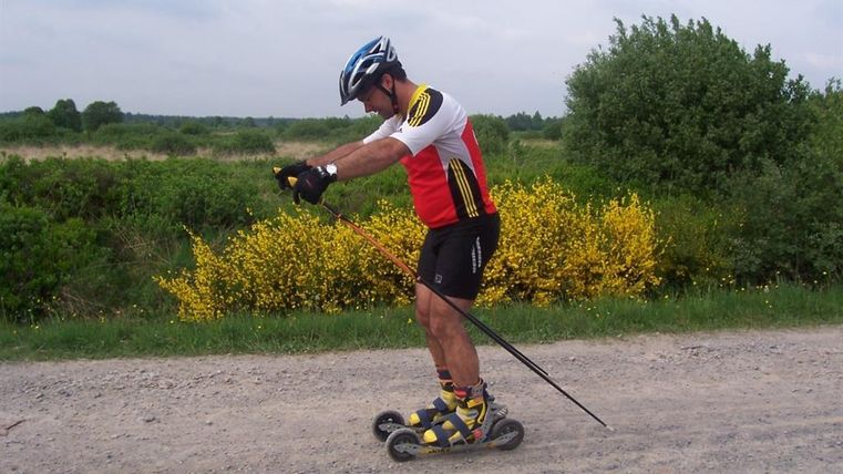 Ein Mann fährt auf einem Rollskiweg und trägt Sportkleidung sowie einen Helm. Im Hintergrund sind grüne Sträucher und gelbe Blumen zu sehen.