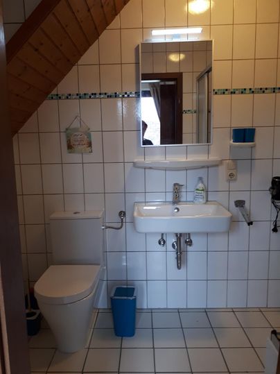 Ein einfaches Badezimmer mit weißen Fliesen und einer Dusche. Es gibt ein Waschbecken, eine Toilette und einen großen Spiegel.