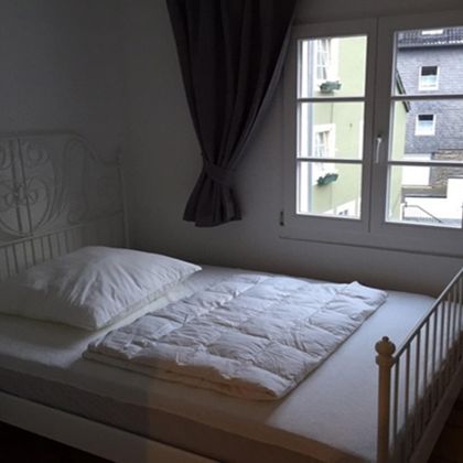 Ein einfaches Schlafzimmer mit einem weißen Bett und einem Fenster. Die Wände sind hell und es gibt Vorhänge, die das Licht sanft dämpfen.