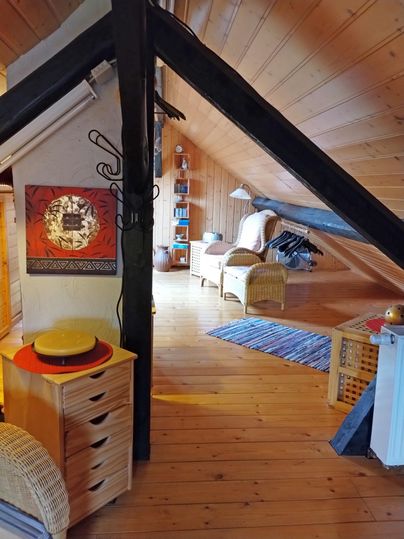 Ein gemütlicher Dachraum mit Holzdecken und einem warmen Ambiente. Die Einrichtung besteht aus einem Korbsessel, Regalen und einem rustikalen Kommode.