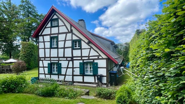 Ein traditionelles Fachwerkhaus mit rotem Dach und grünen Fensterrahmen. Umgeben von einem gepflegten Garten und hohen Hecken.