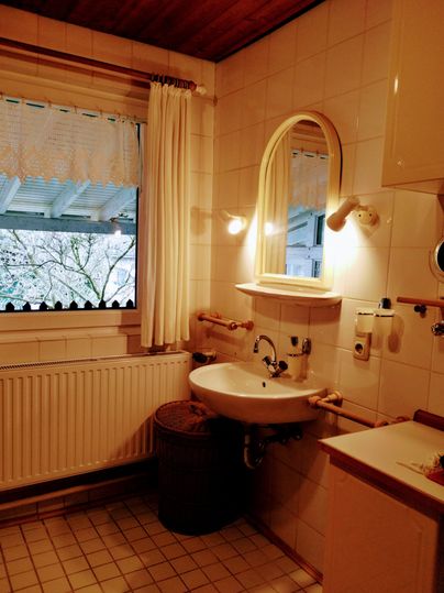 Ein kleines Badezimmer mit einem Waschbecken, einem Spiegel und einem Fenster. Die Wände sind hell gefliest und es gibt eine warme Atmosphäre durch die Beleuchtung.