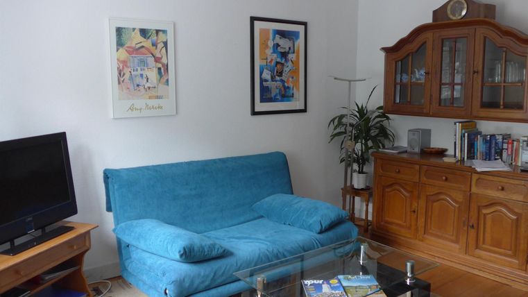 Ein gemütliches Wohnzimmer mit einem blauen Sofa und einem Holzschrank. An der Wand hängen zwei Bilder, und eine Pflanze steht in der Ecke.