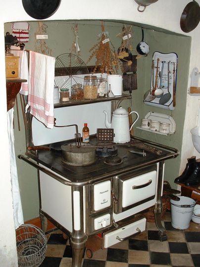 Eine nostalgische Küche mit einem alten Holzofen, Regalen und verschiedenen Küchenutensilien. An der Wand hängen getrocknete Kräuter und weitere Kochutensilien.