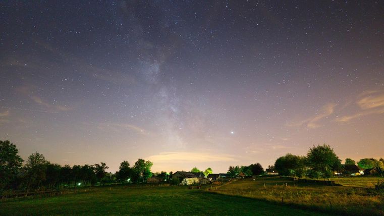 Ein klarer Nachthimmel voller Sterne und die Milchstraße sind sichtbar. Im Vordergrund liegt eine ruhige Landschaft mit Bäumen und sanften Hügeln.