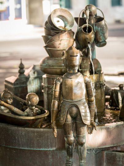 Eine skulpturale Brunneninstallation mit einer metallenen Figur und verschiedenen Gefäßen. Der Brunnen ist kunstvoll gestaltet und vermittelt eine kreative Atmosphäre.