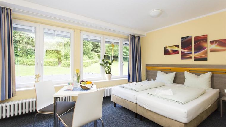 Ein helles Hotelzimmer mit zwei Betten, einem Tisch und Stühlen. Große Fenster sorgen für viel Tageslicht und einen Blick ins Grüne.