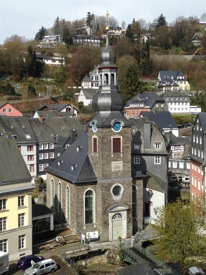 Eine steinerne Kirche mit einem hohen Turm, umgeben von historischen Häusern. Im Hintergrund sind bewaldete Hänge und weitere Gebäude sichtbar.
