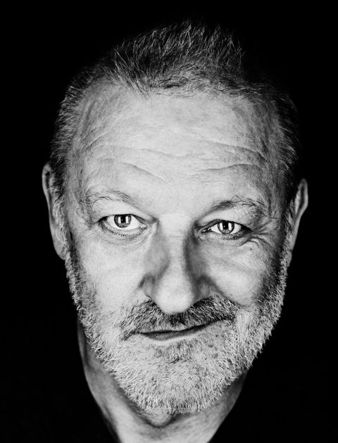Ein Porträt eines lächelnden Mannes mit grauem Bart und kurzen Haaren. Der Hintergrund ist schwarz, was den Fokus auf sein Gesicht legt.