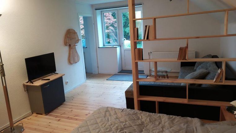 Ein gemütlicher Raum mit einem Fernseher und einem Sofa. Durch ein Fenster fällt Tageslicht und es gibt eine Holztrennwand.